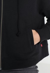 Sudadera con cremallera negra de tela texturizada, con bolsillos delanteros y capucha con cordón. Presenta una pequeña etiqueta de logo roja cerca del bolsillo.