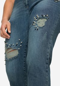 Jeans de mezclilla en azul medio con parches desgastados, adornados con cuentas blancas y negras alrededor de la zona de la rodilla.
