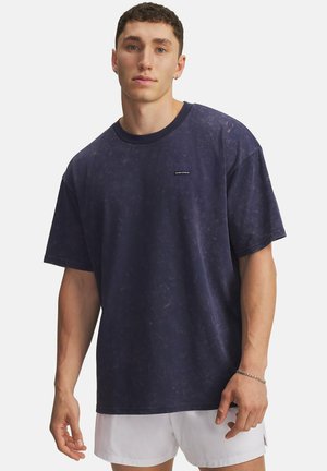 Young man wearing a dark blue short-sleeve t-shirt and white shorts, standing against a plain light background.