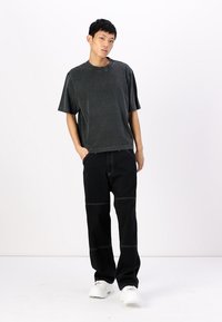 Grijze oversized t-shirt met korte mouwen, relaxte pasvorm, gedragen met zwarte, wijde broek met contrasterende stiksels. Model staat met de handen in de zakken.