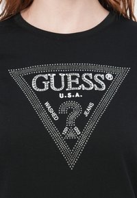 Black cotton t-shirt featuring a triangular rhinestone design with "GUESS U.S.A.", "WASHED", and "JEANS" text, accented with shiny details.