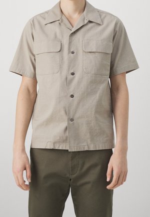 Mężczyzna w beżowej koszuli na krótki rękaw z dwoma kieszeniami na piersi i oliwkowozielonych spodniach, stojący na jednolitym tle.