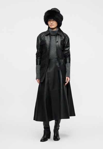 Γυναίκα που φοράει μαύρο γούνινο καπέλο, μαύρο δερμάτινο μπουφάν, μαύρη δερμάτινη φούστα, γκρι πουλόβερ με ζιβάγκο και μαύρα μποτάκια με τακούνι, μπροστά σε απλό φόντο.