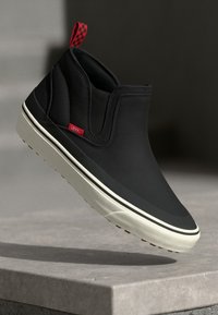 Black slip-on sneaker with a smooth, matte finish. Features red checker-patterned tag and a contrasting white midsole with brown accents.