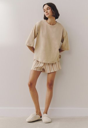Młoda kobieta stoi w pomieszczeniu, ubrana w beżowy, oversize t-shirt, pasiastą krótką spódnicę i białe, puszyste kapcie na tle jednolitej ściany.