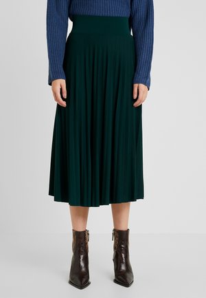 Donkergroene geplooide rok van gemiddelde lengte gedragen met een blauw gebreide trui en bruine enkellaarsjes met slangenhuidprint op een lichtgrijze achtergrond.