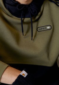 Olivgrön kort sweatshirt med svarta detaljer, dragsko i huvan och en broderad Adidas-logga på framsidan. Texturerad tyg och passform.