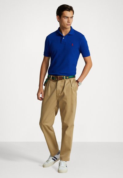 Modrá polo košeľa, khaki plisované nohavice, opasok so zelenými akcentmi, biele tenisky s modrými prúžkami. Neformálny outfit s klasickým strihom.