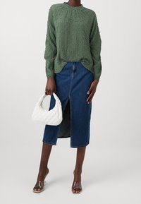 Blouse verte texturée à manches longues, jupe midi en denim avec fente à l'avant, sac à main matelassé blanc et sandales à talons transparentes.