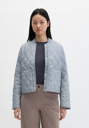 Femme aux longs cheveux noirs portant une veste matelassée gris clair, un haut gris foncé et un pantalon beige, debout devant un fond blanc uni.