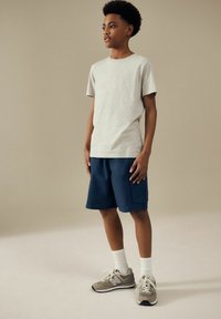 Maglietta grigia a maniche corte abbinata a pantaloni navy. Il modello indossa calze bianche e sneakers grigie con dettagli neri. Outfit semplice e casual.