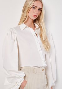 Blusa blanca con cuello estructurado, mangas abullonadas y tapeta de botones. Combinada con pantalones de rayas beige claros y cintura fruncida.
