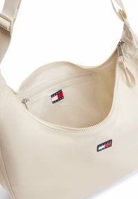 Bolso de hombro de nailon beige con cierre de cremallera y bolsillo interior. Presenta la etiqueta del logo de Tommy Jeans. Textura suave, correa ajustable.