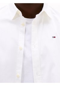 Camicia bianca con bottoni, collo alla francese, bottoni beige e un piccolo logo ricamato in rosso, bianco e blu. Indossata sopra a una t-shirt bianca.