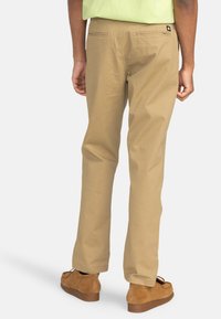 Chinos en coton beige avec jambes droites, poches arrière et une petite étiquette logo sur la ceinture, portés avec des chaussures en daim marron.