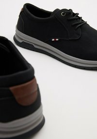 Zapatos sintéticos negros con un acabado texturizado, suela gris, punta redonda y un acento de cuero marrón en el talón. Tienen cordones y un pequeño logotipo.