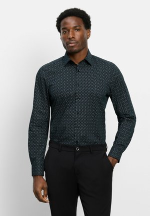 Man die een donker, patroon lang mouw overhemd en zwarte broek draagt, staande met één hand in de zak tegen een effen achtergrond.