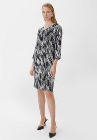 Schwarz-weiß gemustertes Kleid mit V-Ausschnitt und Dreiviertelärmel, aus glattem Stoff, knielang, kombiniert mit schwarzen High Heels.