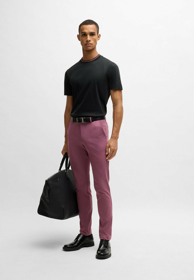 Czarny t-shirt z okrągłym dekoltem, slim-fit, w połączeniu z jasnofioletowymi spodniami. Czarne skórzane buty i duża czarna torba podróżna z uchwytami.