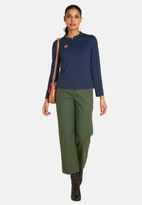 Marineblaues Langarmoberteil mit hohem Ausschnitt und Patch-Details, kombiniert mit hochgeschnittenen grünen Hosen und schwarzen Schnürstiefeln.