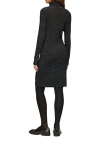 Schwarzes, langärmeliges Kleid mit hohem Kragen, aus strukturiertem Stoff mit metallischen Fylekeln. Kombiniert mit schwarzen, mit Nieten besetzten Schuhen und blickdichten Strumpfhosen.