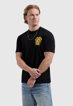 Blonde man met zwart T-shirt met gele leeuw opdruk en lichtblauwe spijkerbroek, kijkt opzij met gekruiste armen voor zich.