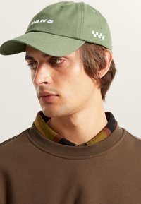 Olivengrøn cap med broderet "VANS"-logo og ternet mønster på siden, parret med en brun sweatshirt og en mønstret skjorte med krave.