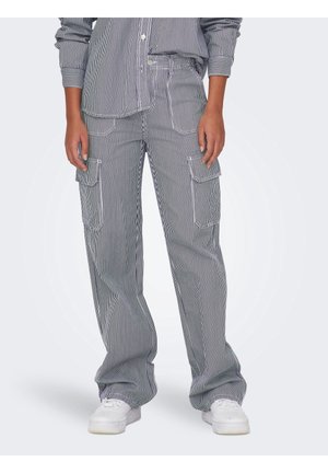 Personne portant un pantalon cargo rayé vertical noir et blanc avec poches plaquées et baskets blanches, debout avec les mains détendues le long du corps.
