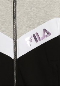 Chaqueta con cremallera de Fila que presenta una sección superior gris, secciones inferiores blancas y negras, y un logo púrpura brillante en el pecho.