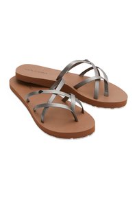 Beige flip-flopit, jossa on ruskea kumipohja ja metallinhohtoinen hopea ristivyö. Pohjassa on kevyt tekstuuri ja brändin nimi "Volcom" on näkyvissä.