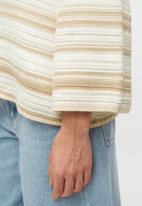 Pull en maille à rayures beige et crème avec des manches larges et un tissu texturé. L'ourlet est arrondi, laissant apparaître une main reposant sur un jean en denim clair.