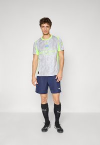 Szürke rövid ujjú sportpóló neon zöld díszítésekkel, "ETIHAD" logóval, sötétkék rövidnadrággal és fekete, térdig érő zoknival, semleges háttéren.