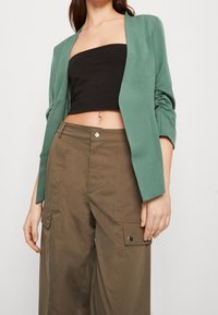 Grön kavaj med en strukturerad yta, öppen front och långa ärmar som bärs med högmidjade bruna cargobyxor med sidofickor.