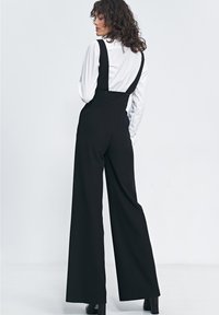 Czarne ogrodniczki o szerokich nogawkach, wykonane z materiału, z regulowanymi ramiączkami, bocznymi kieszeniami i dopasowanym krojem, zestawione z białą koszulą z kołnierzykiem.