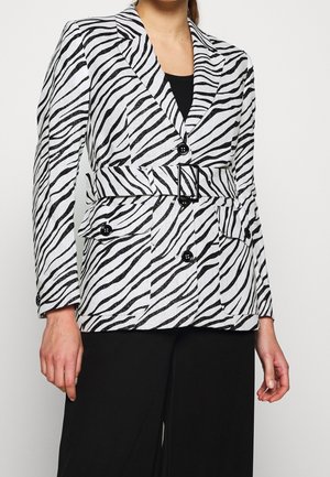 Vrouw draagt een zwart-wit zebraprint blazer met riem en zwarte knopen over een zwarte broek tegen een effen achtergrond.