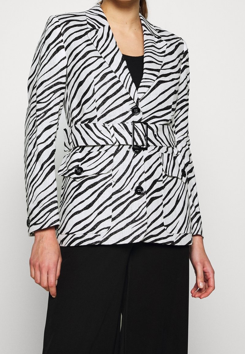 Femme portant un blazer ceinturé à rayures zèbre noir et blanc avec des boutons noirs sur un pantalon noir, sur un fond uni.