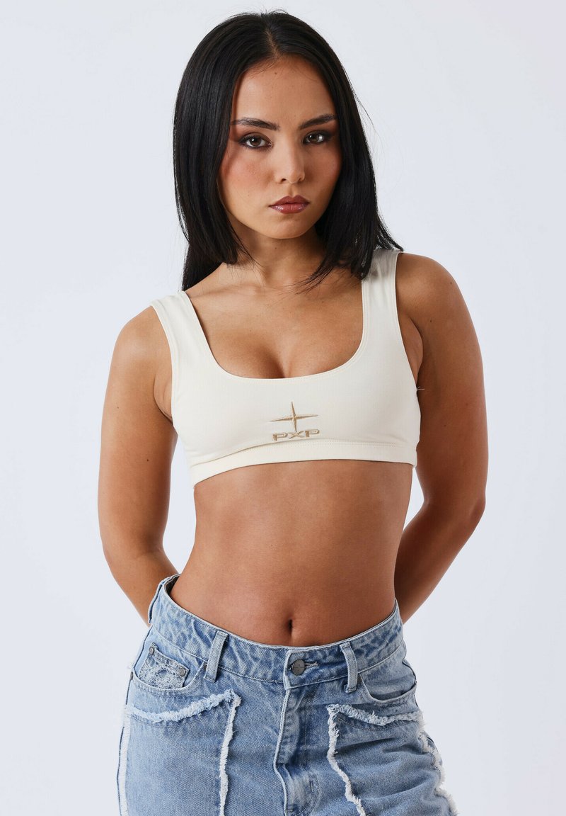 Beige crop top met brede bandjes, van textuurstof, met een gouden "PXP" logo in het midden. Gepaarde met versleten lichtblauwe denim shorts.