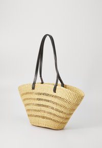 Kudottu olkipussukka avoimella kuvioinnilla, luonnollinen beige väri ja mustat nahkaremmit valkoisella ommella. Kaltevat puolet.