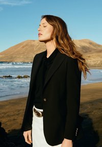 Czarna marynarka z złotymi guzikami, noszona na czarnym topie. Połączona z białymi spodniami i brązowym paskiem. Włosy rozwiane, w tle plaża.