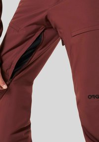 Maroon ski pants with a water-resistant fabric, featuring side zippered pockets, a smooth texture, and a logo accent near the knee.