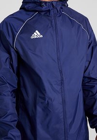 Veste imperméable bleu marine avec capuche, ornée de détails en piping blanc et d'un logo Adidas sur le côté gauche de la poitrine. Fabriquée en matière lisse et légère.