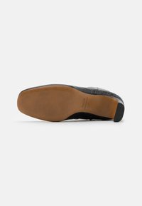Zapato de cuero negro con un patrón texturizado, parecido al de un cocodrilo, que presenta un tacón negro liso y una suela de goma color tan con ranuras horizontales.