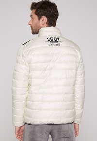 Giacca leggera tipo puffer crema chiaro con cuciture orizzontali, colletto alto e testo grafico nero sul retro. Presenta un orlo a coste con accenti neri.