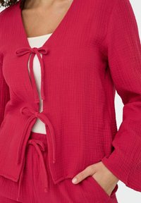 Giacca rosa testurizzata con maniche lunghe, scollatura a V profonda e chiusure a cravatta, abbinata a un fondo dal taglio rilassato.