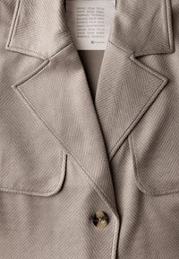 Jasnoszary żakiet z wzorem w rybią kratę, z klapami typu notch oraz dwiema przednimi kieszeniami. Zawiera okrągły guzik i miękką fakturę.