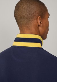 Camisa polo azul marino con cuello a rayas amarillas, que muestra una tela texturizada y un diseño simple y clásico con un acabado limpio en el cuello.