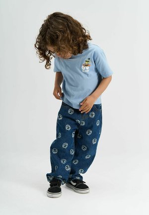 Heled sinine lühikeste varrukatega särk, millel on kiri "MINE AEGLASALT" ja joonistus. Tumedast sinisest teksapüksid, millel on korduv valge logo muster. Mustad spordijalatsid.