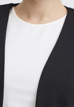 Close-up af en person iført en hvid ribbet rundhalset top under en sort ærmeløs vest.