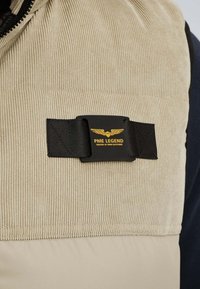 Bežani kordurasti materijal s crnom trakicom i crnim pravokutnim zakrpama na kojoj se nalazi žuti logo s krilima i tekst "PME Legend Tested at High Altitude".