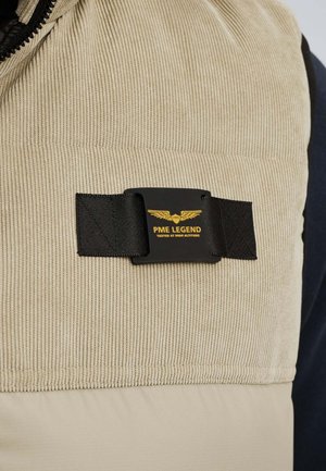 Beige corduroy fabric with a black strap and black rectangular patch featuring yellow wings logo and "PME Legend Tested at High Altitude" text.